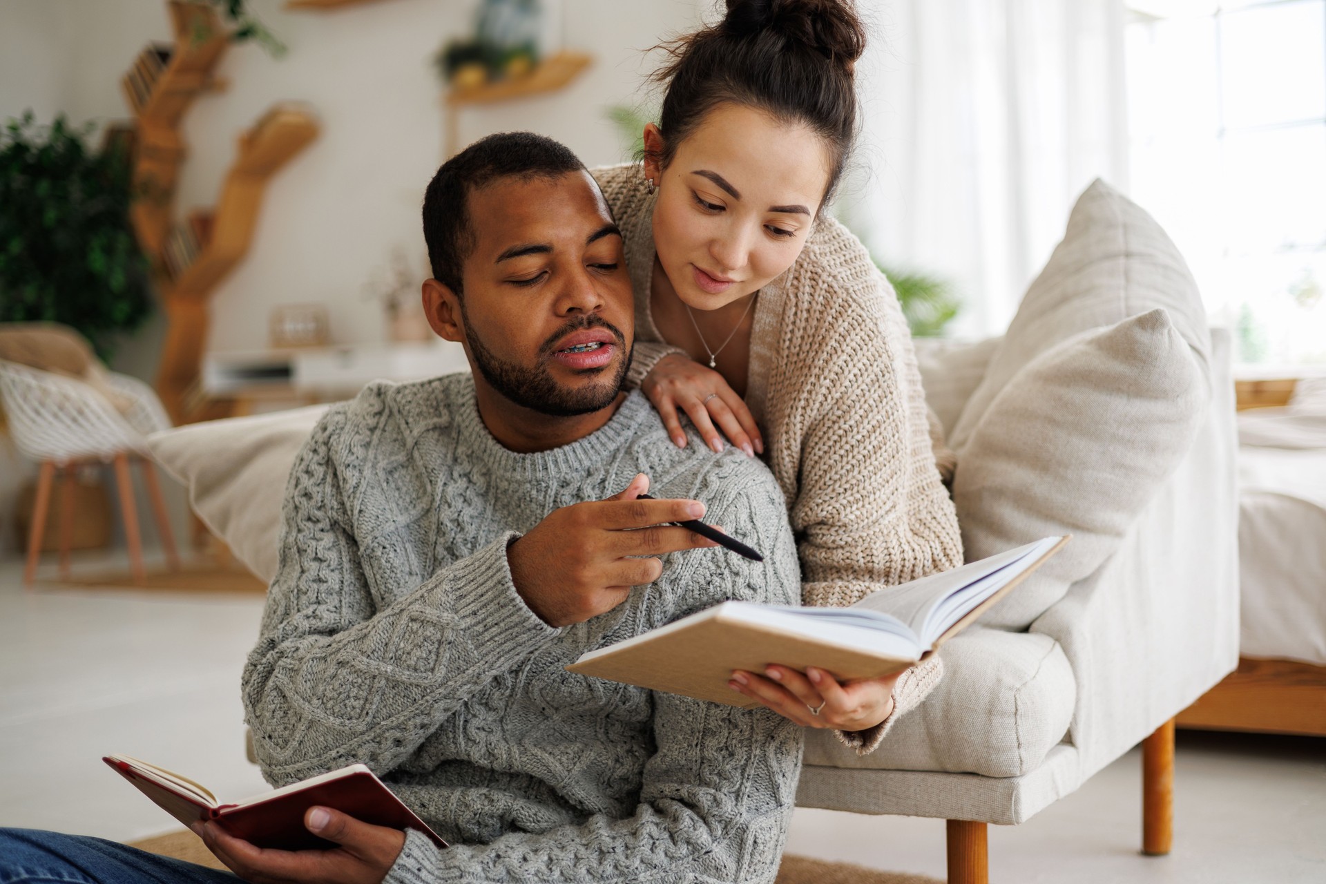 Young romantic multicultural couple in warm clothes reading book together near notebook at home